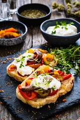 Tasty sandwiches - toasted bread with burrata cheese, anchovies, bell pepper, olives, tomatoes and thyme on wooden table
