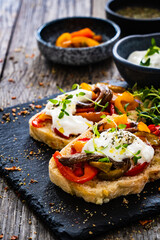 Tasty sandwiches - toasted bread with burrata cheese, anchovies, bell pepper, olives, tomatoes and thyme on wooden table
