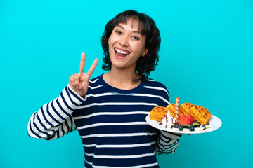 Young Argentinian woman holding waffles isolated on blue background smiling and showing victory sign