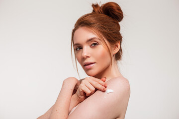 Red-haired beautiful young woman with clean facial skin applies cream on her shoulder in the studio on a white background. Beauty and body skin care
