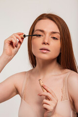 Fototapeta premium Beautiful young redheaded fresh female model with clear skin does makeup and mascara eyelashes in the studio. Beauty