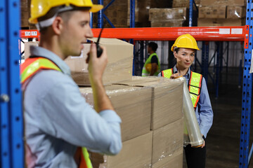 Group of warehouse workers with hardhats and reflective jackets wrapping boxes in stretch film parcel on pallet while control stock and inventory in retail warehouse logistics distribution center