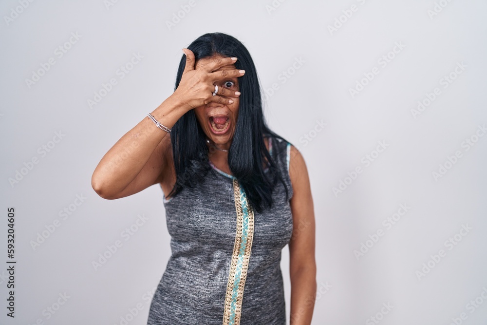 Sticker Mature hispanic woman standing over white background peeking in shock covering face and eyes with hand, looking through fingers with embarrassed expression.