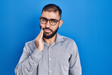 Middle east man with beard standing over blue background touching mouth with hand with painful expression because of toothache or dental illness on teeth. dentist