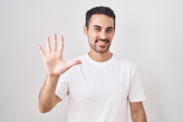 Handsome hispanic man standing over white background showing and pointing up with fingers number five while smiling confident and happy.