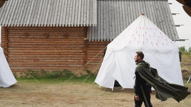 Skillful medieval warrior walks about territory of settlement on cloudy day. Wind blows around long cloak of young bearded knight side view