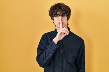 Young man wearing glasses over yellow background asking to be quiet with finger on lips. silence and secret concept.