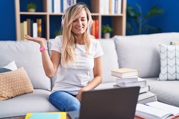 Obraz premium Young blonde woman studying using computer laptop at home smiling cheerful presenting and pointing with palm of hand looking at the camera.