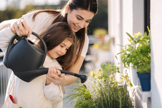 Happy daughter watering plants with her beautiful mom. Girl's in bright overall gardening on balcony