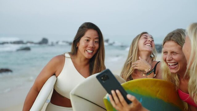Happy multiracial surfer girls in bikini taking selfies holding surfboards on beach then looking at phone screen. Sexy beautiful young surfing women in swimwear take selfie photographs after session.