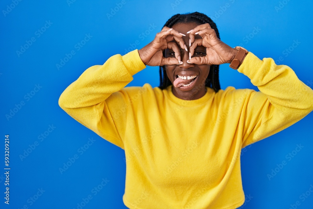 Wall mural Beautiful black woman standing over blue background doing ok gesture like binoculars sticking tongue out, eyes looking through fingers. crazy expression.