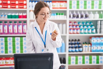 Young blonde woman pharmacist using computer holding pills bottle at pharmacy