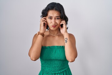 Young hispanic woman standing over isolated background covering ears with fingers with annoyed expression for the noise of loud music. deaf concept.
