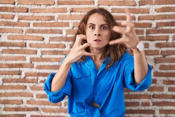 Beautiful brunette woman standing over bricks wall shouting frustrated with rage, hands trying to strangle, yelling mad