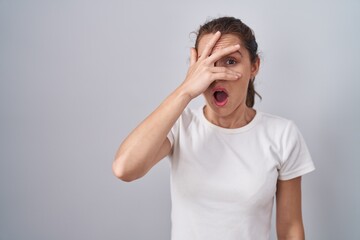 Beautiful brunette woman standing over isolated background peeking in shock covering face and eyes...