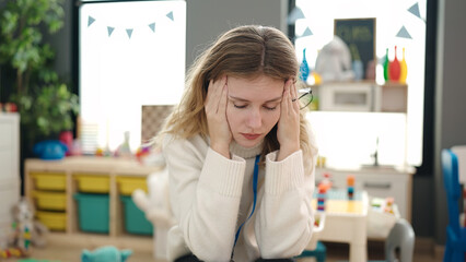 Young blonde woman preschool teacher stressed sitting on chair at kindergarten