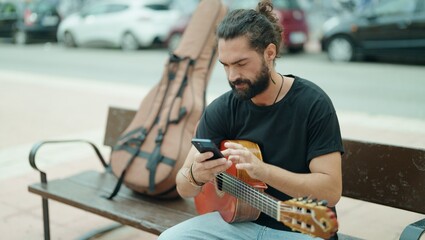 Young hispanic man musician holding classical guitar using smartphone at street