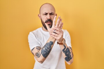 Young hispanic man with tattoos standing over yellow background suffering pain on hands and...