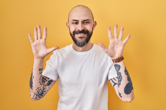 Young hispanic man with tattoos standing over yellow background showing and pointing up with fingers number ten while smiling confident and happy.