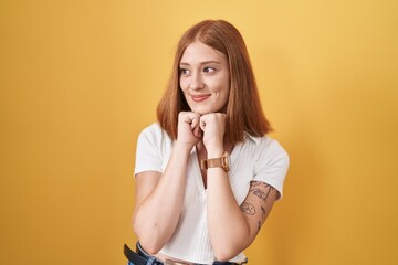 Young redhead woman standing over yellow background laughing nervous and excited with hands on chin...