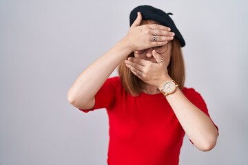 Young redhead woman standing wearing glasses and beret covering eyes and mouth with hands, surprised and shocked. hiding emotion