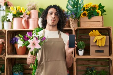 Hispanic man with curly hair working at florist shop showing smartphone screen making fish face with mouth and squinting eyes, crazy and comical.