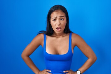 Fototapeta na wymiar Hispanic woman standing over blue background in shock face, looking skeptical and sarcastic, surprised with open mouth