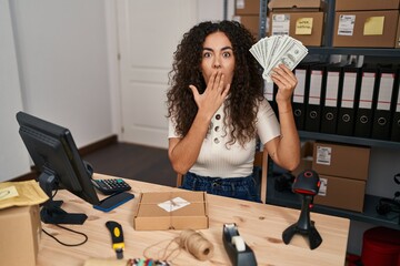 Young hispanic woman working at small business ecommerce holding dollars covering mouth with hand, shocked and afraid for mistake. surprised expression