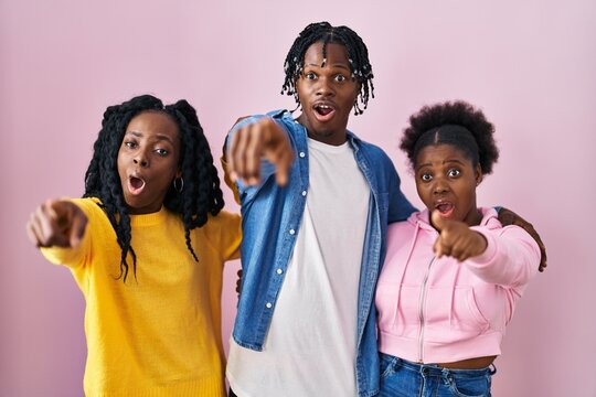 Group of three young black people standing together over pink background pointing with finger surprised ahead, open mouth amazed expression, something on the front