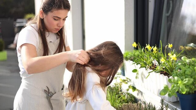 Beautiful young mom helps her daughter to get dressed overall for gardening on balcony. Adorable girl smile