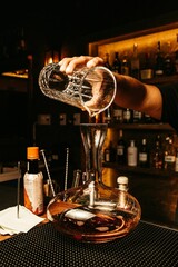 Vertical of a barman making cocktail in the big glass jar with alcoholic drinks in the bakcground