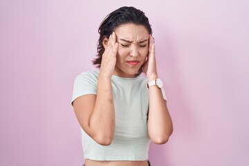 Hispanic young woman standing over pink background with hand on head, headache because stress. suffering migraine.