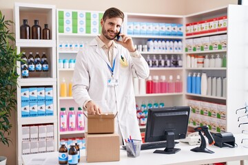 Young caucasian man pharmacist talking on smartphone holding pills bottle at pharmacy