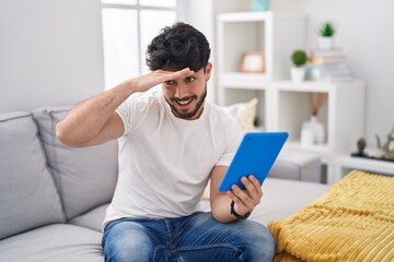 Hispanic man with beard using touchpad sitting on the sofa very happy and smiling looking far away with hand over head. searching concept.