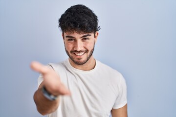 Hispanic man with beard standing over white background smiling cheerful offering palm hand giving assistance and acceptance.
