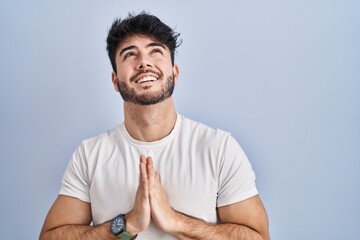 Hispanic man with beard standing over white background begging and praying with hands together with...