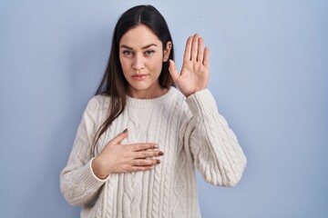Young brunette woman standing over blue background swearing with hand on chest and open palm,...