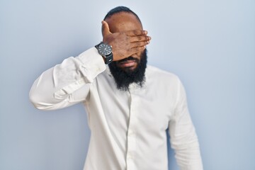 Obraz premium African american man standing over blue background covering eyes with hand, looking serious and sad. sightless, hiding and rejection concept