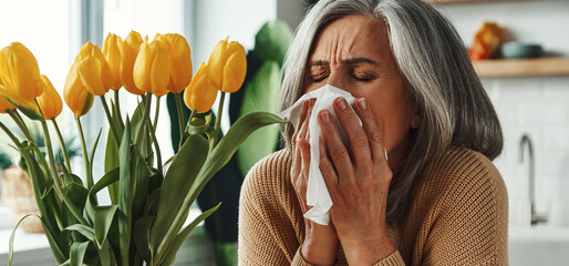 Senior woman sneezing an keeping eyes closed while sitting near the bunch of tulips at home