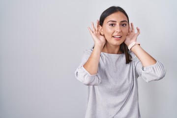 Young hispanic woman standing over white background trying to hear both hands on ear gesture, curious for gossip. hearing problem, deaf