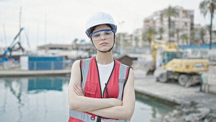 Young beautiful hispanic woman builder standing with serious expression and arms crossed gesture at seaside
