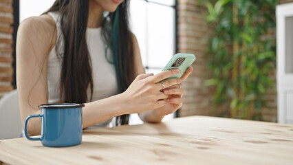 Young beautiful hispanic woman using smartphone drinking coffee at dinning room