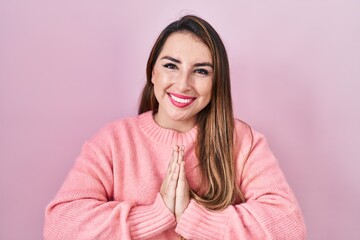 Fototapeta premium Young hispanic woman standing over pink background praying with hands together asking for forgiveness smiling confident.