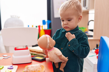 Adorable caucasian boy standing with relaxed expression playing with baby doll at kindergarten