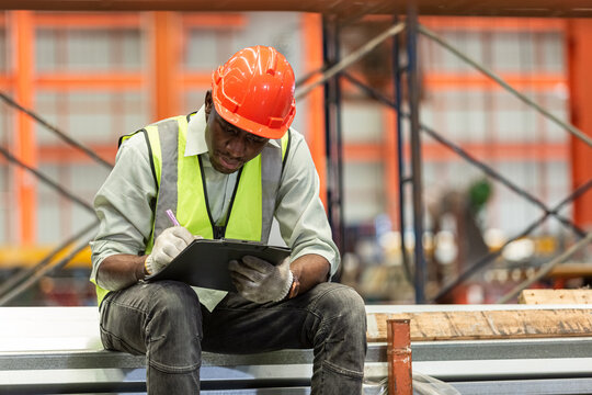 Happy african american workers wear red helmet working take notes on paperwork clipboard sitting in factory. Heavy Industrial manufacturing workplace.