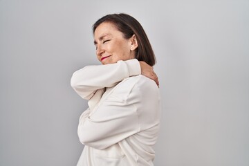 Middle age hispanic woman standing over isolated background hugging oneself happy and positive,...
