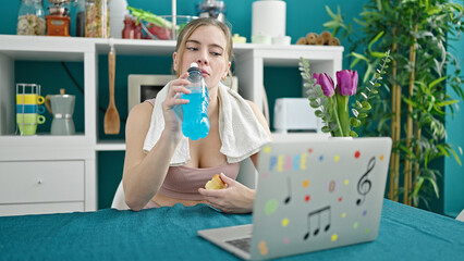 Young blonde woman wearing sportswear using laptop drinking energetic beverage at dinning room
