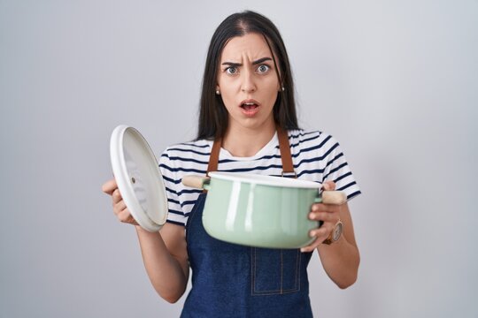 Young brunette woman wearing apron holding cooking pot in shock face, looking skeptical and sarcastic, surprised with open mouth