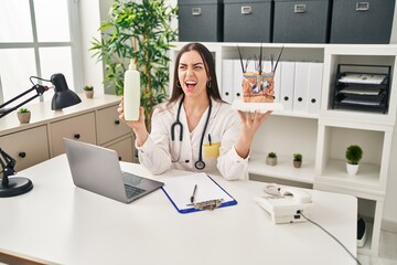 Hispanic doctor woman holding model of human anatomical skin and hair angry and mad screaming frustrated and furious, shouting with anger. rage and aggressive concept.