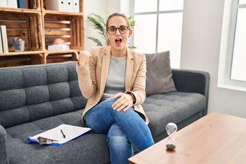 Fototapeta premium Young woman working at consultation office surprised pointing with hand finger to the side, open mouth amazed expression.